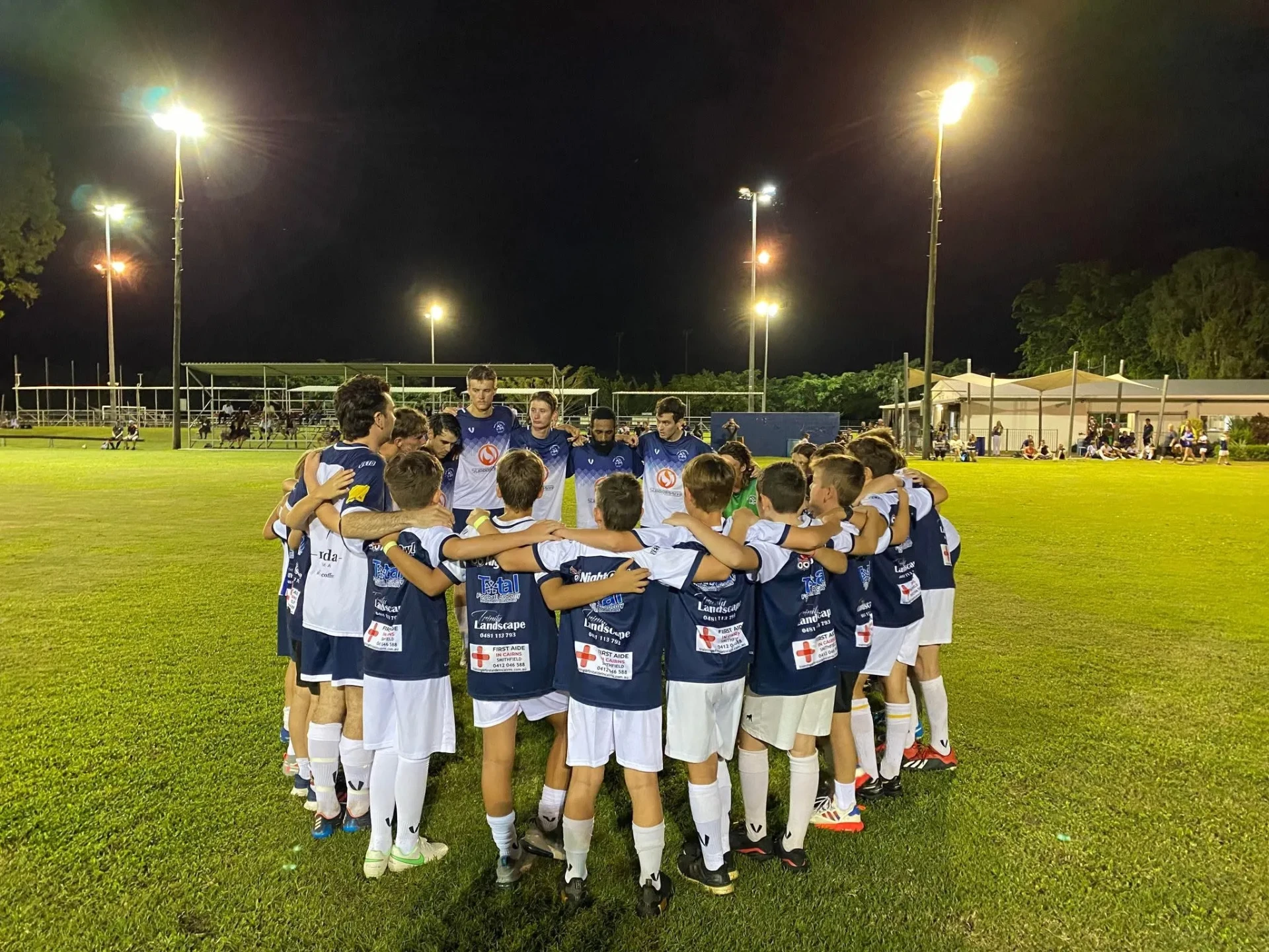Junior and senior players in a huddle on the pitch under floodlights at Pennell Field